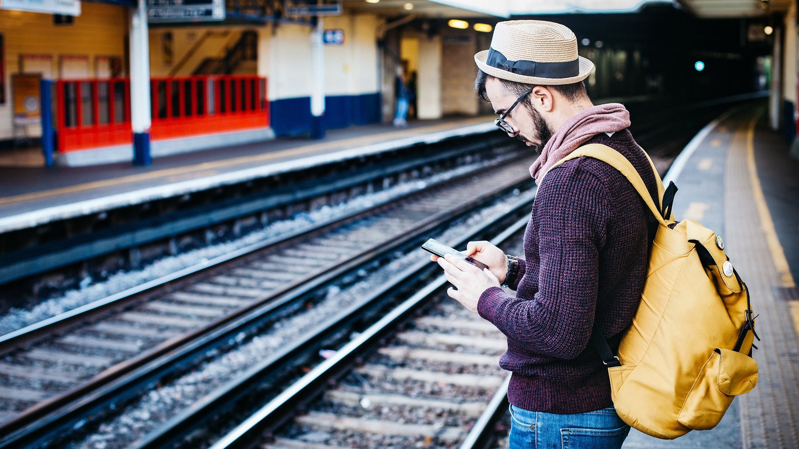 Traveller wearing a yellow backpack and checking a phone while waiting on a railway platform, illustrating a carry-on backpack for a 3-day trip