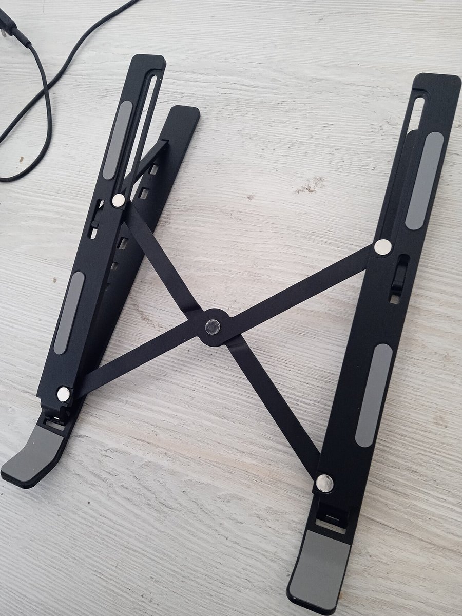 Black folding aluminium laptop stand with an X-shaped frame, rubber pads and front lips, opened on a wooden table